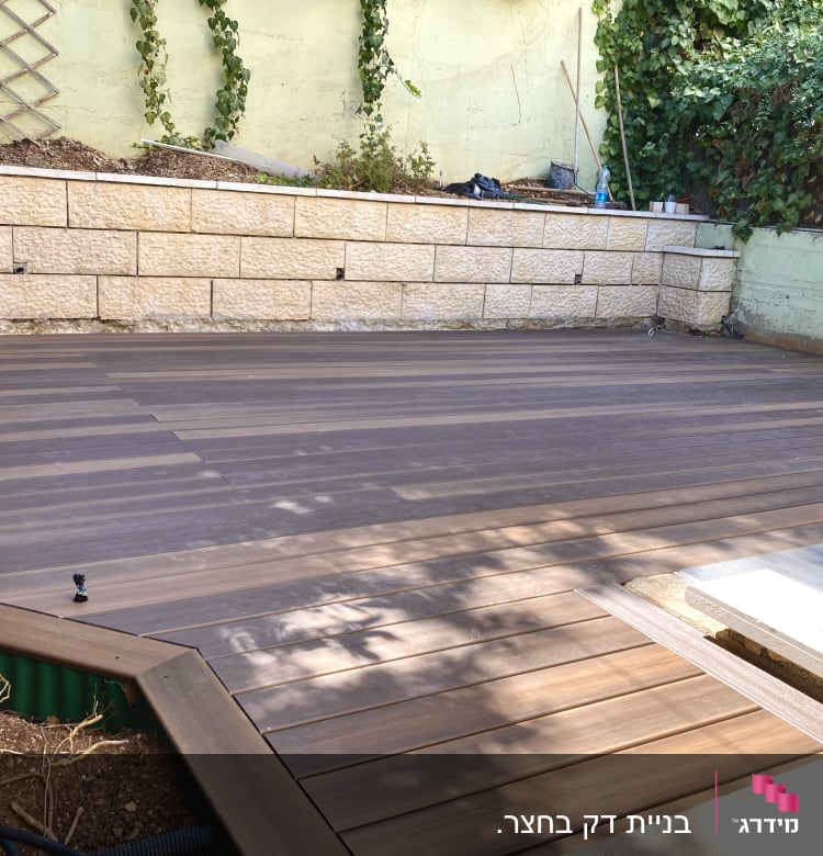 דק עץ בגינה עם קיר אבן וצמחייה מסביב